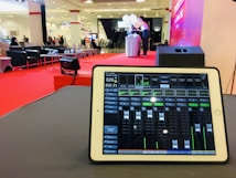 A tablet is displaying audio mixing software on a table, with a large hall visible in the background. The room is set up for an event, with red carpet flooring, seating arrangements, and a stage area where two people stand next to a table with balloons.