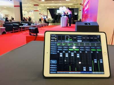 A tablet is displaying audio mixing software on a table, with a large hall visible in the background. The room is set up for an event, with red carpet flooring, seating arrangements, and a stage area where two people stand next to a table with balloons.