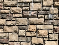 A close-up view of a stone wall constructed with rectangular and irregularly shaped stones in various shades of beige and brown. The stones are arranged in a staggered pattern, creating a textured surface that highlights the natural variations and rough surface of each stone.
