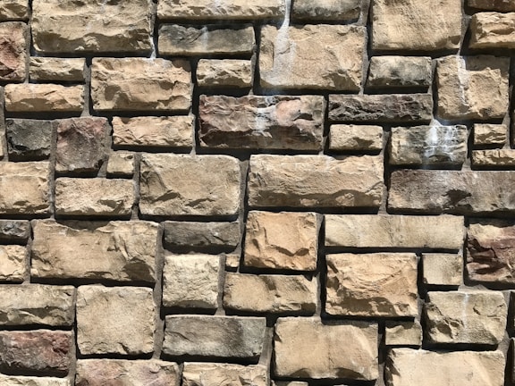 A close-up view of a stone wall constructed with rectangular and irregularly shaped stones in various shades of beige and brown. The stones are arranged in a staggered pattern, creating a textured surface that highlights the natural variations and rough surface of each stone.