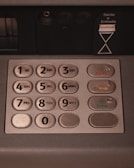 A close-up view of a numeric keypad typically found on ATMs, featuring metal buttons numbered 0 to 9, along with function keys labeled in another language. The panel includes instructions for envelope deposit.