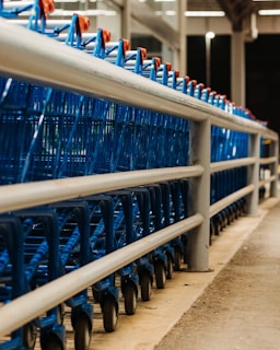 A line of blue shopping carts is neatly arranged behind a metal railing in what appears to be a supermarket or store setting. The perspective shows the carts from a low angle, highlighting their wheels and metal frames.