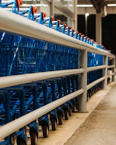 A line of blue shopping carts is neatly arranged behind a metal railing in what appears to be a supermarket or store setting. The perspective shows the carts from a low angle, highlighting their wheels and metal frames.