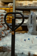 The image captures a close-up view of a pallet jack's handle in what appears to be a warehouse or storage area. Surrounding the pallet jack are multiple stacks of packaged goods wrapped in plastic, likely bags of flour or similar products. Shelving units filled with various items are visible in the background.