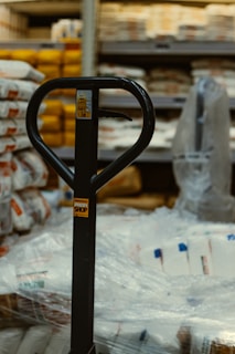 The image captures a close-up view of a pallet jack's handle in what appears to be a warehouse or storage area. Surrounding the pallet jack are multiple stacks of packaged goods wrapped in plastic, likely bags of flour or similar products. Shelving units filled with various items are visible in the background.