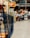 A shopping cart is visible in the foreground with a store setting in the blurred background. Shelves lined with various products can be seen, and people appear to be moving around, contributing to a busy atmosphere.