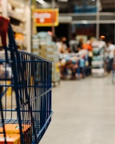 A shopping cart is visible in the foreground with a store setting in the blurred background. Shelves lined with various products can be seen, and people appear to be moving around, contributing to a busy atmosphere.