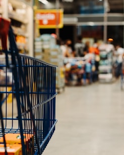 A shopping cart is visible in the foreground with a store setting in the blurred background. Shelves lined with various products can be seen, and people appear to be moving around, contributing to a busy atmosphere.