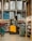 A warehouse or large store interior with shelves stacked high with packaged goods and supplies. A forklift with a yellow and black design is positioned in the center aisle, suggesting an industrial setting. The shelves hold products like large bags of rice or flour, boxed items, and various merchandise. Brightly colored cleaning tools are displayed on the left side, while more packaged goods fill the shelves to the right.