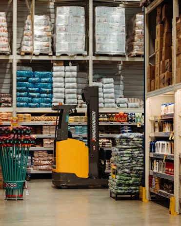 A warehouse or large store interior with shelves stacked high with packaged goods and supplies. A forklift with a yellow and black design is positioned in the center aisle, suggesting an industrial setting. The shelves hold products like large bags of rice or flour, boxed items, and various merchandise. Brightly colored cleaning tools are displayed on the left side, while more packaged goods fill the shelves to the right.