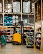 A warehouse or large store interior with shelves stacked high with packaged goods and supplies. A forklift with a yellow and black design is positioned in the center aisle, suggesting an industrial setting. The shelves hold products like large bags of rice or flour, boxed items, and various merchandise. Brightly colored cleaning tools are displayed on the left side, while more packaged goods fill the shelves to the right.