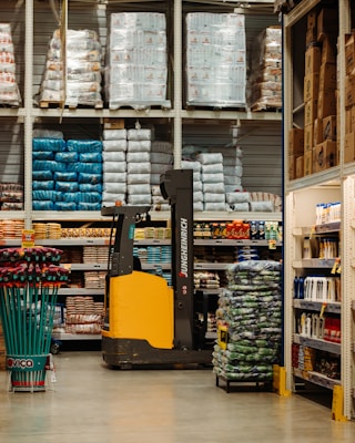 A warehouse or large store interior with shelves stacked high with packaged goods and supplies. A forklift with a yellow and black design is positioned in the center aisle, suggesting an industrial setting. The shelves hold products like large bags of rice or flour, boxed items, and various merchandise. Brightly colored cleaning tools are displayed on the left side, while more packaged goods fill the shelves to the right.