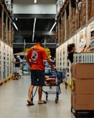 A man wearing an orange shirt and sandals pushes a shopping cart down an aisle in a large warehouse-style store. Another person wearing dark clothing and a face mask is reaching for items on a shelf. The aisle is lined with well-stocked shelves and boxes, and other shoppers are visible in the background.