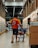 A man wearing an orange shirt and sandals pushes a shopping cart down an aisle in a large warehouse-style store. Another person wearing dark clothing and a face mask is reaching for items on a shelf. The aisle is lined with well-stocked shelves and boxes, and other shoppers are visible in the background.