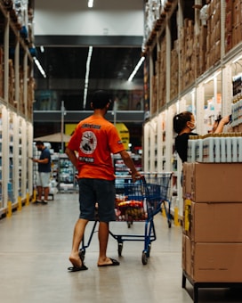 A man wearing an orange shirt and sandals pushes a shopping cart down an aisle in a large warehouse-style store. Another person wearing dark clothing and a face mask is reaching for items on a shelf. The aisle is lined with well-stocked shelves and boxes, and other shoppers are visible in the background.
