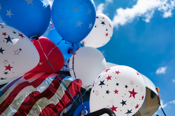 Balloons with star patterns in red, white, and blue colors float against a bright blue sky. The scene includes flag-themed decorations suggesting a celebratory or patriotic event.