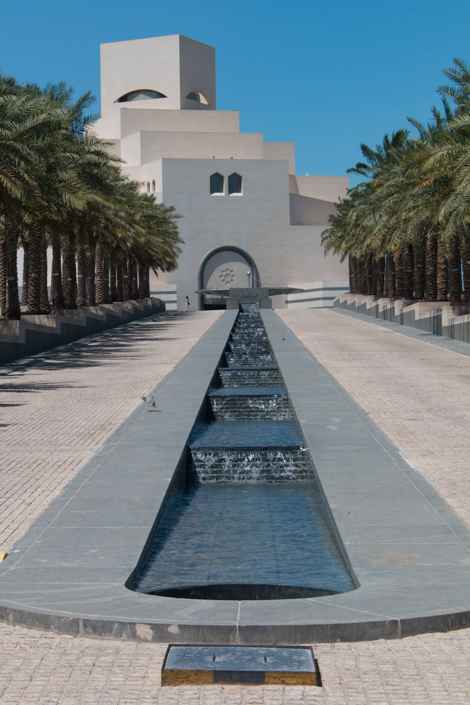 The image depicts a large, modern building with a curved roof and multiple levels, situated on a street lined with palm trees. The building features a central entrance flanked by two arches, creating an architectural focal point. A long, rectangular pool is the centerpiece of the scene, surrounded by several steps leading up to it. The clear blue sky provides a serene backdrop for this urban landscape.