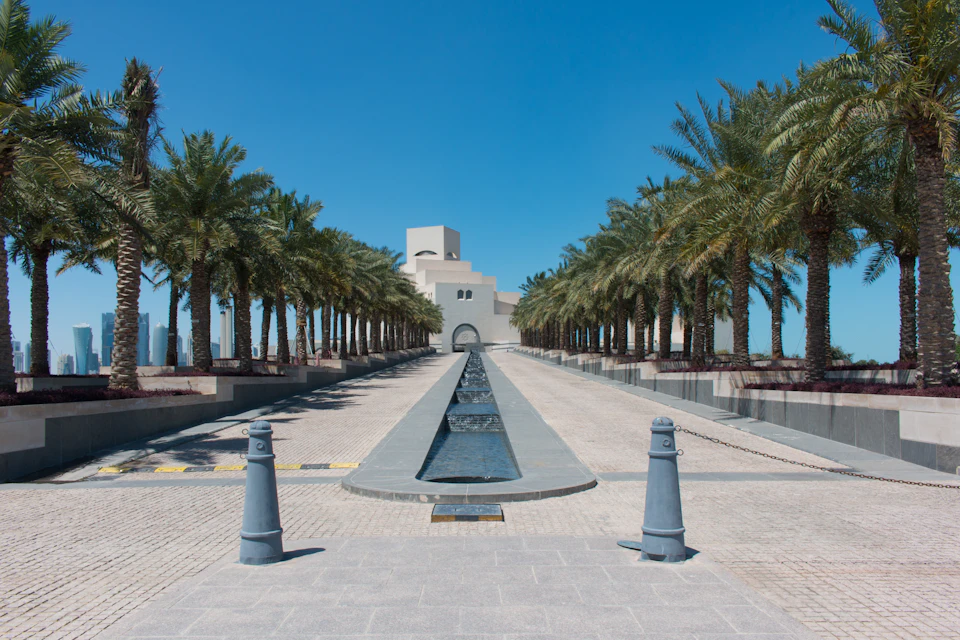 The image depicts a serene scene at the Dubai Museum, featuring a long, straight walkway flanked by tall palm trees on both sides. The walkway is lined with gray stone columns and leads towards a large fountain situated in the center. In the distance, buildings can be seen, suggesting that this location is within or near the city of Dubai.