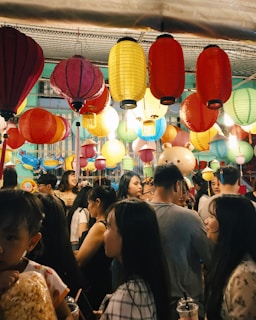 A bustling scene at a festival or celebration features a crowd of people under a canopy of vibrant, multicolored lanterns. The lanterns hang in various colors including red, yellow, and blue, illuminating the lively atmosphere. People of different ages mingle, some holding snacks and drinks.