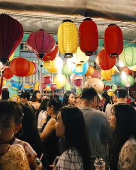 A bustling scene at a festival or celebration features a crowd of people under a canopy of vibrant, multicolored lanterns. The lanterns hang in various colors including red, yellow, and blue, illuminating the lively atmosphere. People of different ages mingle, some holding snacks and drinks.