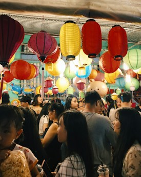 A bustling scene at a festival or celebration features a crowd of people under a canopy of vibrant, multicolored lanterns. The lanterns hang in various colors including red, yellow, and blue, illuminating the lively atmosphere. People of different ages mingle, some holding snacks and drinks.
