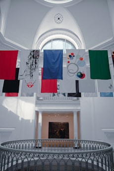 A series of colorful banners or flags hang inside a large, bright gallery space featuring an ornate ceiling with a decorative circular pattern. Below is a gallery with white columns and a black and white photograph or painting on the wall.