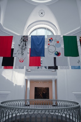 A series of colorful banners or flags hang inside a large, bright gallery space featuring an ornate ceiling with a decorative circular pattern. Below is a gallery with white columns and a black and white photograph or painting on the wall.