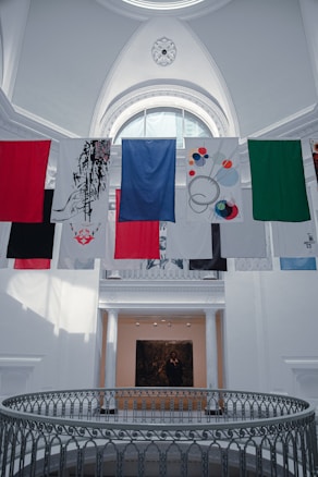 A series of colorful banners or flags hang inside a large, bright gallery space featuring an ornate ceiling with a decorative circular pattern. Below is a gallery with white columns and a black and white photograph or painting on the wall.