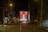 A nighttime urban scene with a deserted street flanked by tall buildings. In the background, a large digital billboard displays a bright advertisement featuring text and a partially visible figure. The street is dimly lit with streetlights, casting reflections on the wet pavement, while a single vehicle is visible driving down the road.