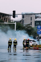 Firefighters in yellow helmets and dark uniforms are attending to a situation on a city street. Smoke is billowing around them as they work near a white car. The scene includes several buildings, a traffic light, road signs, and scattered traffic cones.
