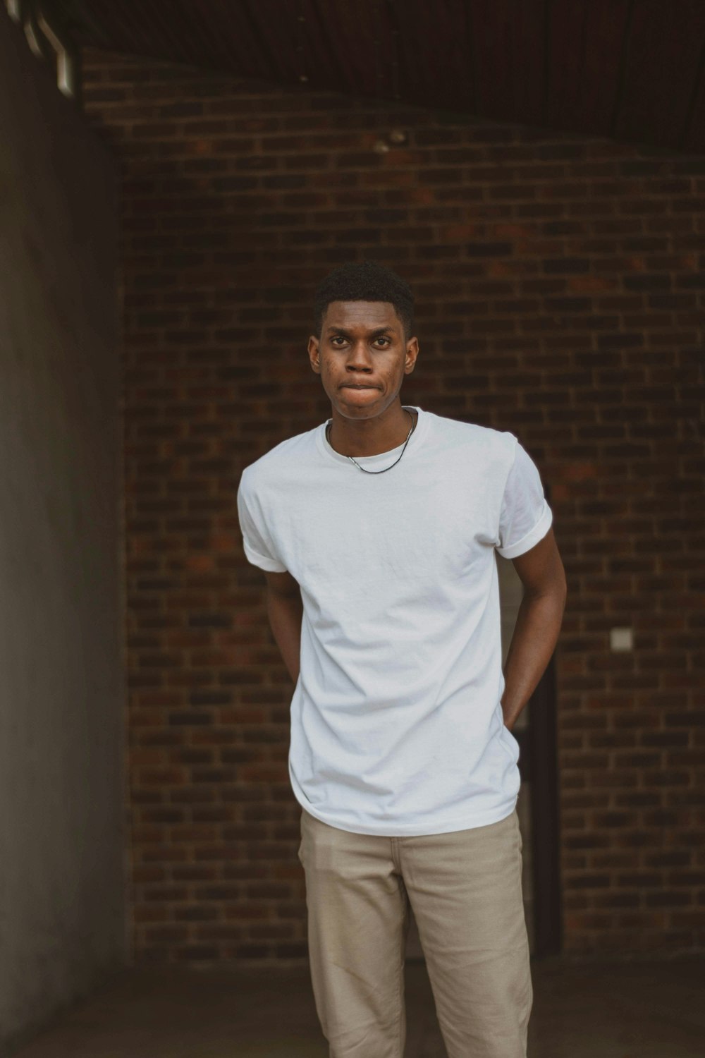 man in white crew neck t-shirt and brown pants standing beside brown brick wall