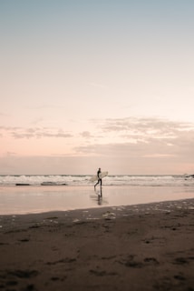 Sunrise over Taghazout beach with a lone surfer carrying a board towards gentle waves
