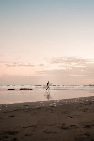 A young surfer carrying a colorful surfboard walking along a sandy beach at sunrise.