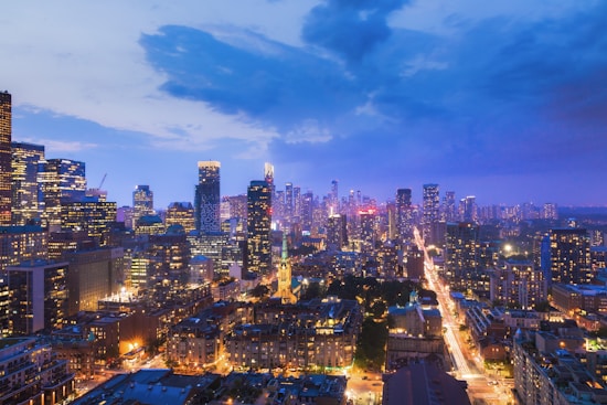 A vibrant cityscape at dusk, featuring numerous skyscrapers illuminated with lights against a deep blue and purple sky. The urban landscape is densely packed with buildings, and a major road with bright light trails cuts through the scene, indicating traffic movement.