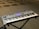 A digital keyboard is placed on a black X-style stand. The keyboard has multiple control knobs, buttons, and a display screen, indicating it is an electronic synthesizer. The background shows several chairs on a carpeted floor, suggesting a music studio or rehearsal space.