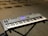 A digital keyboard is placed on a black X-style stand. The keyboard has multiple control knobs, buttons, and a display screen, indicating it is an electronic synthesizer. The background shows several chairs on a carpeted floor, suggesting a music studio or rehearsal space.