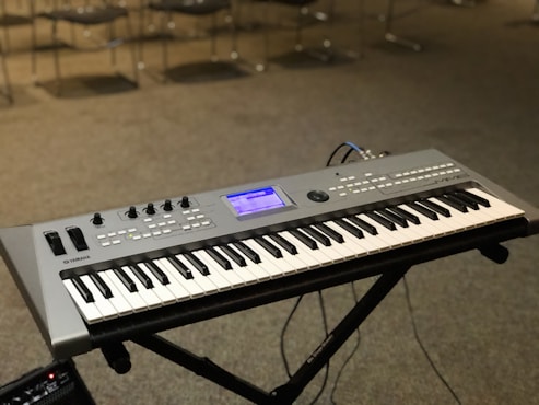A digital keyboard is placed on a black X-style stand. The keyboard has multiple control knobs, buttons, and a display screen, indicating it is an electronic synthesizer. The background shows several chairs on a carpeted floor, suggesting a music studio or rehearsal space.