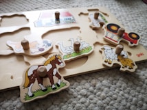 A children's wooden puzzle features various farm animals and a tractor. Each piece has a small wooden handle for easy gripping. The animals depicted include a horse, a group of geese, a pair of sheep, and a cat playing with yarn.