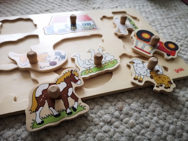 A children's wooden puzzle features various farm animals and a tractor. Each piece has a small wooden handle for easy gripping. The animals depicted include a horse, a group of geese, a pair of sheep, and a cat playing with yarn.