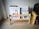 A small wooden bookshelf holds several children's books with colorful covers. The titles are visible and include illustrations of animals and children. The shelf is set against a plain wall and is placed on a light wooden floor, adjacent to a wooden piece of furniture that resembles a chest.
