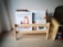 A small wooden bookshelf holds several children's books with colorful covers. The titles are visible and include illustrations of animals and children. The shelf is set against a plain wall and is placed on a light wooden floor, adjacent to a wooden piece of furniture that resembles a chest.