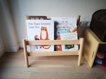 A small wooden bookshelf holds several children's books with colorful covers. The titles are visible and include illustrations of animals and children. The shelf is set against a plain wall and is placed on a light wooden floor, adjacent to a wooden piece of furniture that resembles a chest.