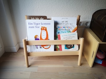 A small wooden bookshelf holds several children's books with colorful covers. The titles are visible and include illustrations of animals and children. The shelf is set against a plain wall and is placed on a light wooden floor, adjacent to a wooden piece of furniture that resembles a chest.