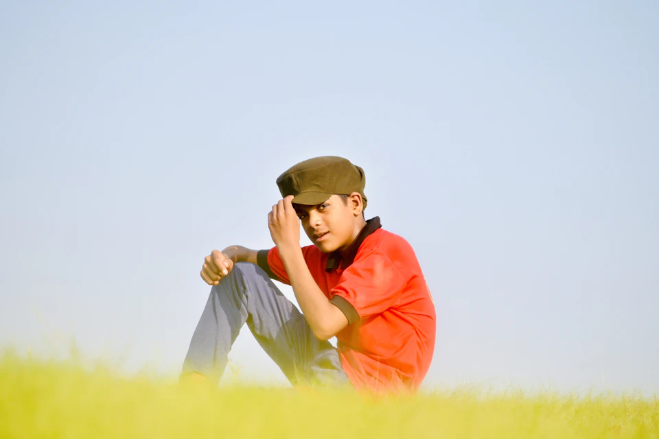 In the image, a young boy is sitting on a grassy field under a clear blue sky. He's wearing an orange shirt and a brown cap that matches his pants. The boy has his hands resting on his knees, suggesting he might be taking a break or simply enjoying the view.