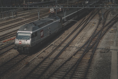 A sleek, modern train positioned on a network of railway tracks under an overcast sky. The train features a silver metallic exterior with SF logos, surrounded by multiple branching tracks that create a complex pattern. Overhead wires and signals are visible, contributing to the industrial setting.