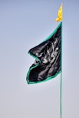 A flag with black fabric and a green border is shown against a clear blue sky. Arabic script in white is visible on the flag, and a small yellow pennant adorns the top of the flagpole.