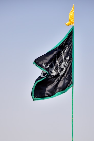 A flag with black fabric and a green border is shown against a clear blue sky. Arabic script in white is visible on the flag, and a small yellow pennant adorns the top of the flagpole.