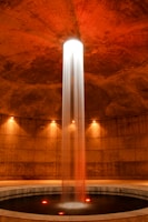 A cylindrical room featuring a central column of water illuminated dramatically from above. The water cascades into a circular pool surrounded by concrete walls. Warm lighting accentuates the reddish-brown tones of the space, adding a serene and mysterious atmosphere.