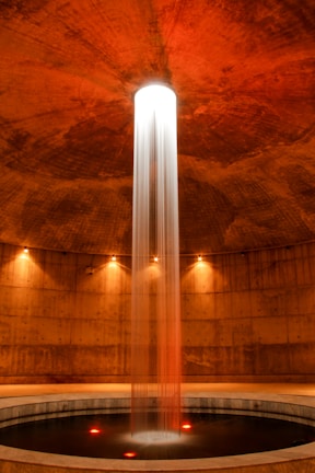 A cylindrical room featuring a central column of water illuminated dramatically from above. The water cascades into a circular pool surrounded by concrete walls. Warm lighting accentuates the reddish-brown tones of the space, adding a serene and mysterious atmosphere.