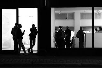 A black and white photograph depicts a group of people standing both inside and outside a building with large glass windows. Inside, a group of people are seen in an art gallery or exhibition space, engaged in viewing the displays. Outside, on the left side of the image, two people are standing and conversing under dim street lighting. The overall scene conveys an urban night-time atmosphere, with the interior of the gallery brightly illuminated, providing a stark contrast to the darker exterior.